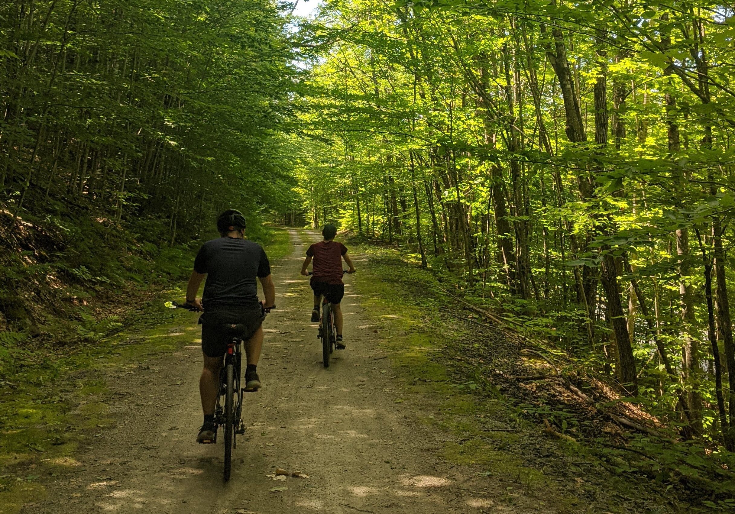 A photo of two people riding bikes on the Northern Rail Trail on a sunny day. Greenery surrounds.
